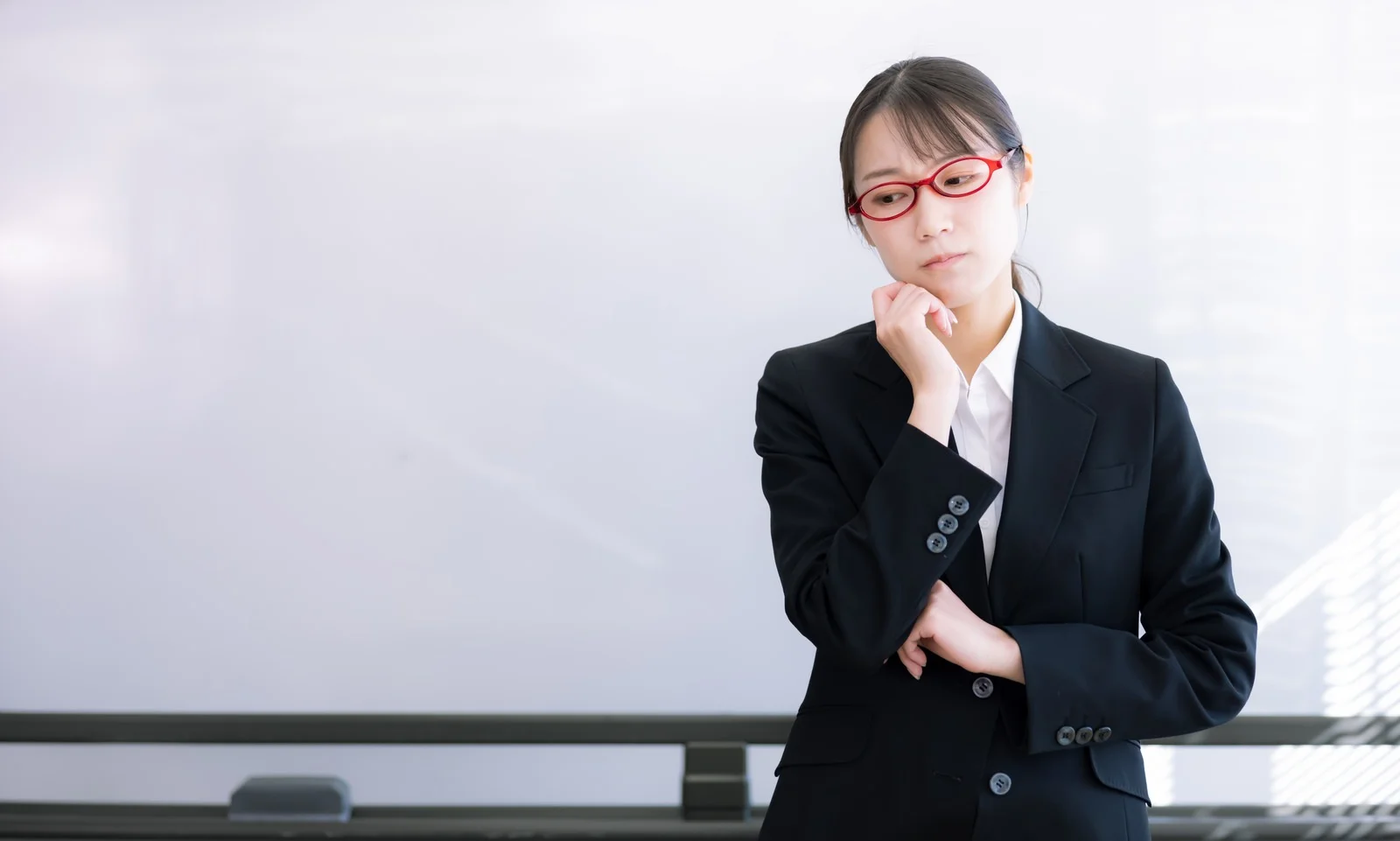 困惑する赤い眼鏡の女性指導員の写真素材です。黒いスーツを着た女性が赤い眼鏡をかけ、ホワイトボードの前で手を顎に当て困った表情を浮かべ、疑問や悩みを表現しています。